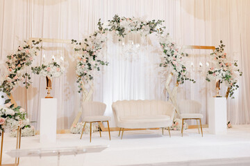 Beautiful decor for the wedding ceremony in the form of a sofa and white flowers on the wedding arch. White furniture in the form of a sofa and armchairs for the bride and groom