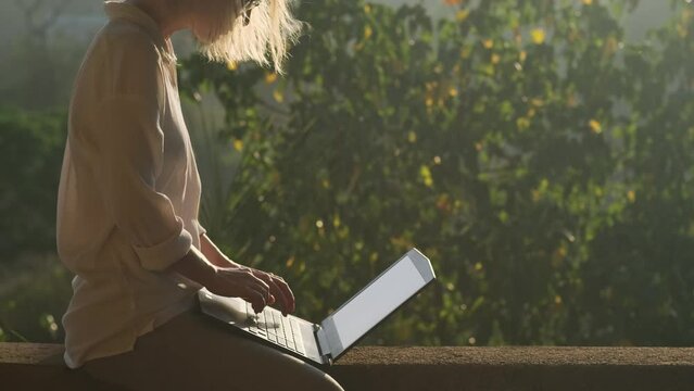 Close-up shot of a young woman typing on a laptop keyboard in sunshine outdoor. Female writer working on the rooftop at sunrise or sunset.