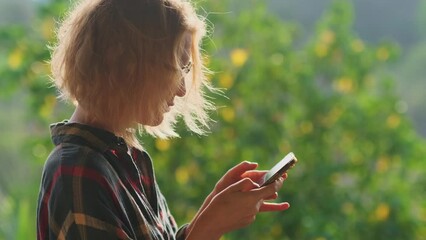 Middle-aged blonde woman using smartphone on the blurred nature background. Female hipster typing text message on the mobile phone. Advertising for mobile technology.