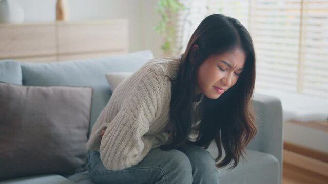 Sick young Asian woman suffering from stomach ache sitting on sofa in living room at home, Abdominal pain, gastritis, and painful periods concept.