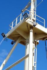 Forward mast of vessel with ladder and platform where are CCTV surveillance camera and led light projectors. Mast has cream color and behind is blue sky as background.