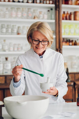 A happy senior pharmacist is standing in a vintage apothecary and adding an ingredient to a mortar...