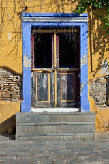 Beautiful entrance with old wooden door, blue to purple lintel, yellow wall and cement staircase during warm afternoon light, in Eleftheroupoli town