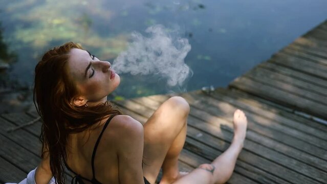 Young sexy red head woman smoking vaper at park near lake in summer holidays.