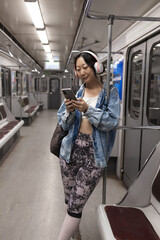 young beautiful and happy Asian Korean woman listening to music with headphones and mobile phone in subway train smiling cheerful in urban lifestyle