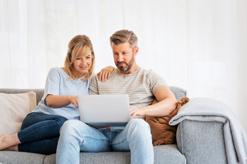 Middle-aged couple wearing casual clothes and sitting at home on the sofa and using a laptop