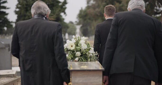 Men, coffin and pallbearers walking at cemetery ceremony outdoor at burial tomb. Death, grief and group casket at funeral, carry to graveyard and family service of people mourning at windy event.