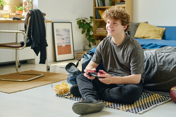 Teenage boy with curly hair sitting on the floor by bed and playing video game while looking at...
