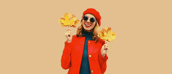 Autumn color style outfit, portrait of beautiful smiling young woman with yellow maple leaves wearing red french beret hat, coat on brown studio background