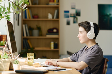 Serious teenage girl listening to music in headphones while sitting by desk in front of computer monitor, looking at screen and networking