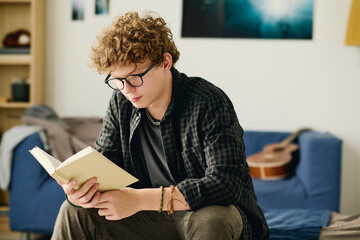 Serious male teenager in eyeglasses reading curious novel while sitting on bed in front of camera...