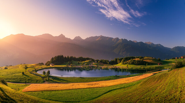 The Wolfsee lake in Fiss (Tirol, Austria) at sunrise between grass, pine trees and a wheat field , in the background the Austrian Alps.