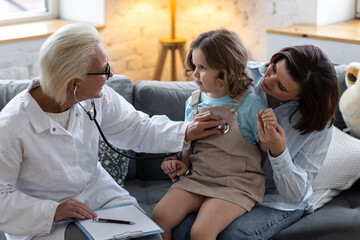 Concept healthcare medical assistance, insurance. Kind senior female paediatrician doctor examining little child sitting on mother's laps, during home visit or clinic check up. Prescribing treatment