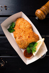Fried breaded pork fillet with lemon, parsley, spices and sauce in a paper box on a dark wooden table.