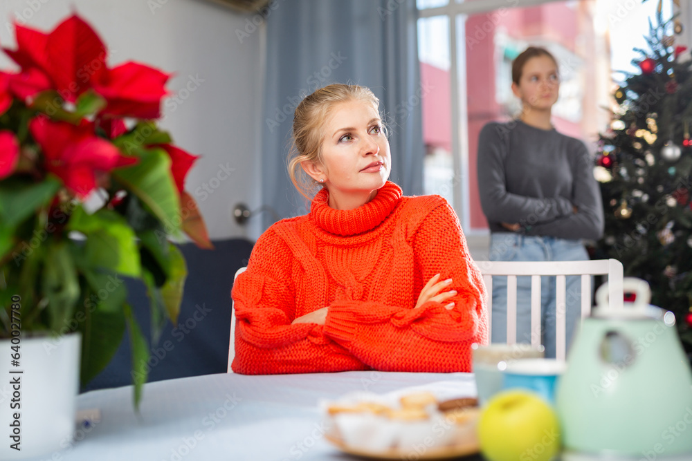 Poster upset mom sitting at table after quarreling with daughter during christmas time