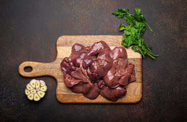 Raw chicken liver on wooden cutting board top view on dark rustic concrete background kitchen table with parsley and garlic. Healthy food ingredient, source of iron, folate, vitamins and minerals