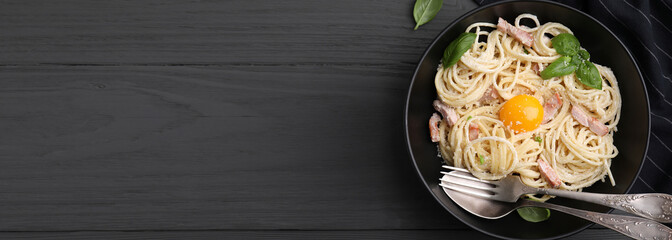 Bowl of delicious pasta Carbonara with egg yolk and cutlery on black wooden table, top view. Banner design with space for text