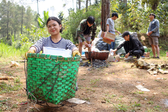 Young Asian girl showing to camera a bag full of trash while friends collecting garbage on the background. Environment support and green nature concept