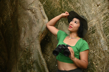 Pretty Asian woman Holding binoculars at the roots of a large tree and are delighted happily. This tree is very large and is a popular attraction for Thai tourists.