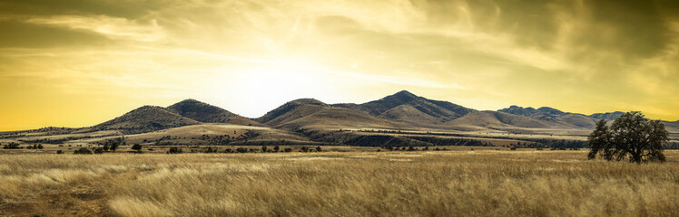 Panorama of a distant mountain range with hills covered in grass in the forground with a dramatic sunset in Arizona.