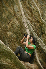 Pretty Asian woman watching through binoculars at huge tree roots. This tree is very large and is a popular tourist destination for Thai tourists.