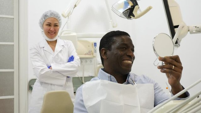 African-american man patient sitting on dental chair and smiling. Asian woman dentist standing in background.