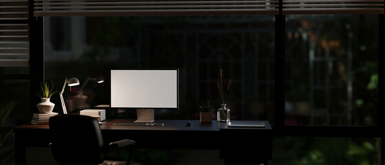 A modern office room at night with light from a table lamp, a computer mockup on a desk