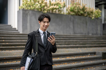 A handsome Asian businessman is checking messages on his phone while walking down the stairs