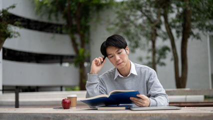 A smart and focused Asian male university student is reading a book in a campus park.