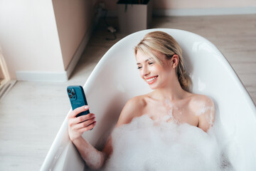 Young beautiful woman having fun while lying in bathtub full of foam at home. Charming smiling model relaxing in luxury bath interior. Female holding smartphone, looking at cellphone screen, use apps