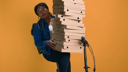 Youthful black woman holding stack of pizza boxes for delivering to a celebration event in neighborhood. African american delivery person cautiously balancing huge order for food delivery.
