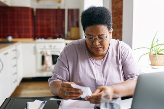 60-years-old black plus size woman pensioner in glasses reading paycheck for electricity or gas feeling upset, examining attentively bill sitting in front of laptop at kitchen table