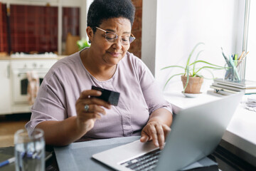 Pretty plus size elderly female of african ethnicity using laptop sitting at kitchen table next to...