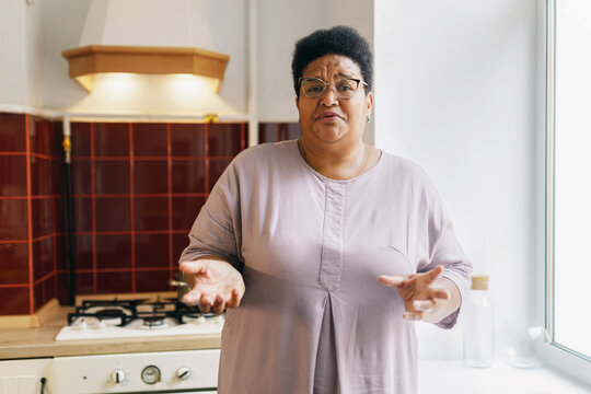 Indoor image of mature housewife or grandma of african ethnicity standing against kitchen gas stove with puzzled facial expression, shrugging and gesticulating, having no idea what to cook for dinner