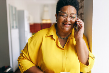 Happy smiling senior black plus size female in yellow shirt and glasses having nice conversation on phone with her best friend, smiling and laughing at jokes, sharing news and rumors - 640106380