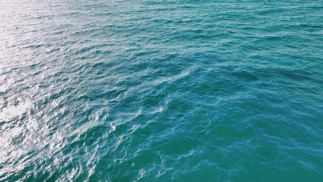 From above we fly over the turquoise sea. Rippling ocean waves. Aerial video of blue sea water background and sun reflection. Water surface waves. Drone flying over the ocean.