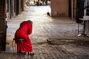 Red dressed tired Moroccan elderly woman taking a rest on a stool placed on an alley inside the...