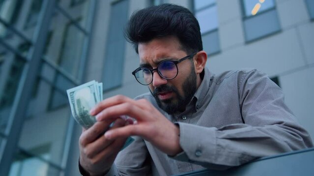 Arabian Indian upset worried man businessman in city outdoors counting money in despair miserable savings tight salary sad guy count dollars cash problem of poverty financial crisis global inflation