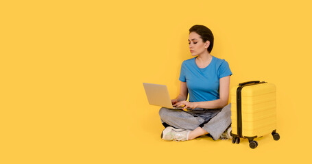 serious woman with yellow suitcase using laptop while sitting with legs crossed on yellow background. Travel enjoy concept