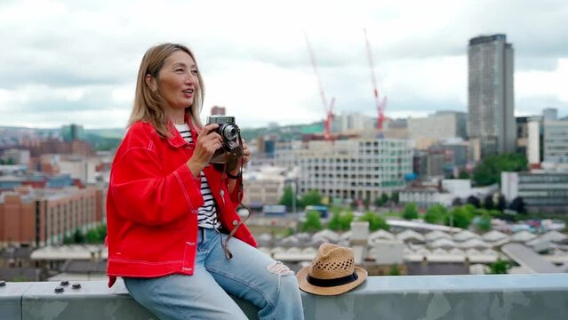 happy asian woman in a red jacket sitting in front of the city Sheffield  and take photos on great summer day