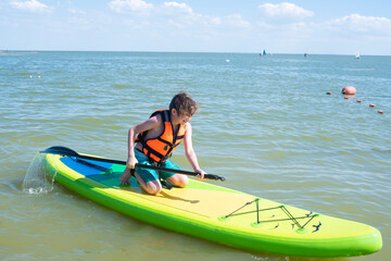 A 10 year old boy is learning to ride a SUP board in the sea. a child in an orange life jacket rides a sapboard alone on the sea on a summer vacation on a sunny day.