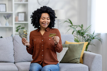 Young happy African American woman holding looking at credit card and holding cash money and bills...