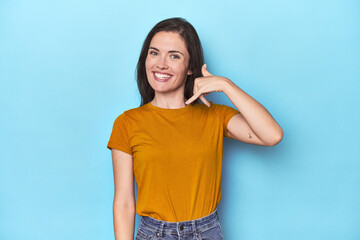 Young caucasian woman on blue backdrop showing a mobile phone call gesture with fingers.
