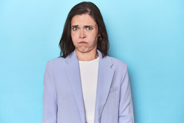 Woman in blue blazer on blue background blows cheeks, has tired expression. Facial expression...