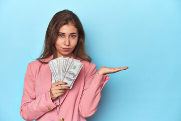 Teen girl in pink, flaunting a handful of dollar bills showing a copy space on a palm and holding another hand on waist.