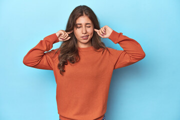 Teen girl in vibrant orange sweater on blue covering ears with fingers, stressed and desperate by a...