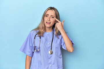 Young caucasian nurse in blue studio showing a disappointment gesture with forefinger.