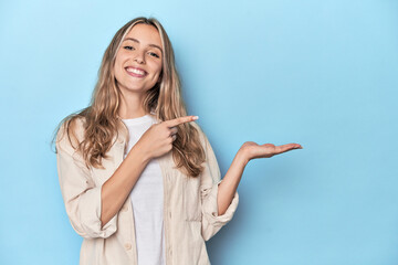 Blonde young caucasian woman in blue studio excited holding a copy space on palm.