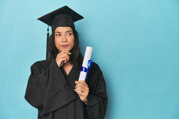Filipina graduate with diploma on blue studio looking sideways with doubtful and skeptical expression.