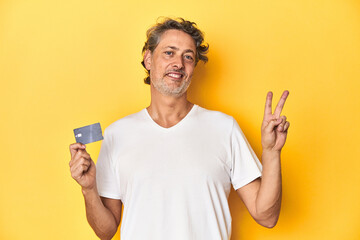 Man holding a credit card, yellow studio backdrop joyful and carefree showing a peace symbol with...
